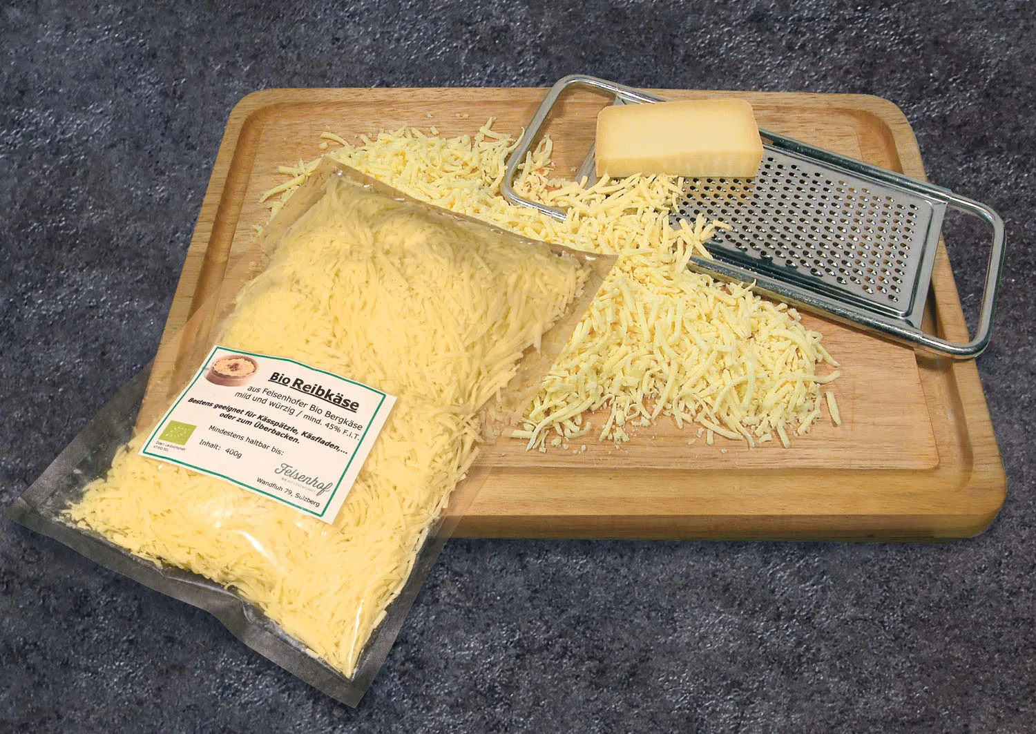 Beutel Bio Reibkäse neben geriebenem Käse und einer Reibe auf einem Holzbrett